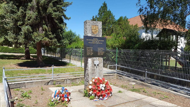 Ein Denkmal für die Opfer der Tschernobyl-Katastrophe, umgeben von einem Metallzaun, Kränzen, Gras, Pflanzen, Bäumen und einem Haus mit Dach und Fenstern, unter einem bewölkten Himmel.