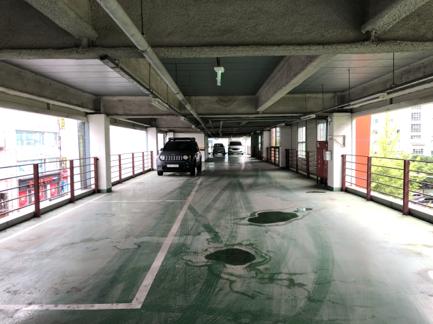 Ein mehrgeschossiges Parkhaus mit parkenden Autos, Gel├Ąndern, Pfeilern, Deckenleuchten, freiliegenden Rohren und umgebenden B├Ąumen, vor einer Kulisse von Geb├Ąuden mit zahlreichen Fenstern in einer Stadtlandschaft.