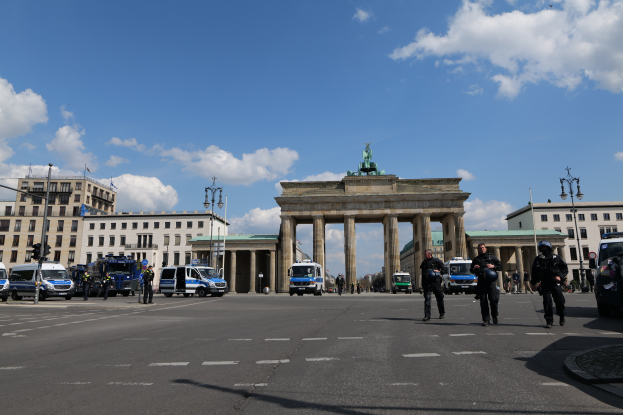 Eine Gruppe von Polizeibeamten vor dem Brandenburger Tor in Berlin, Deutschland, das mit Säulen und einer Statue geschmückt ist, umgeben von Gebäuden mit Fenstern, Fahrzeugen auf der Straße, Laternenmasten und Ampelstangen mit Masten, unter einem bewölkten Himmel.