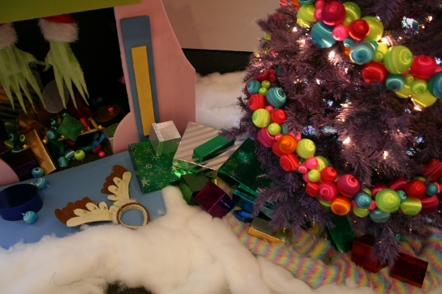 Eine Indoor-Szene mit einem geschm├╝ckten Weihnachtsbaum rechts, Geschenkboxen und Deko in der Mitte und einer H├╝tte mit Deko oben links, mit W├Âlldecke und Schafwolle unten.