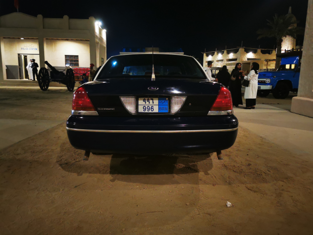 Ein Polizeiwagen parkt nachts vor einem Gebäude, umgeben von einer Gruppe von Menschen, einige halten Kameras, mit einer Kanone im Vordergrund und Gebäuden, Lichtern, Bäumen und einem sternenklaren Himmel im Hintergrund.