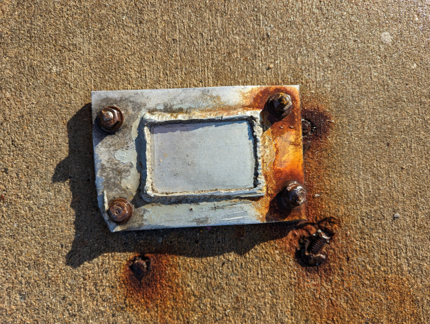 Ein verrostetes Metallschild am Boden mit ein paar verstreuten Schrauben, das Anzeichen von längerer Witterungseinwirkung zeigt.